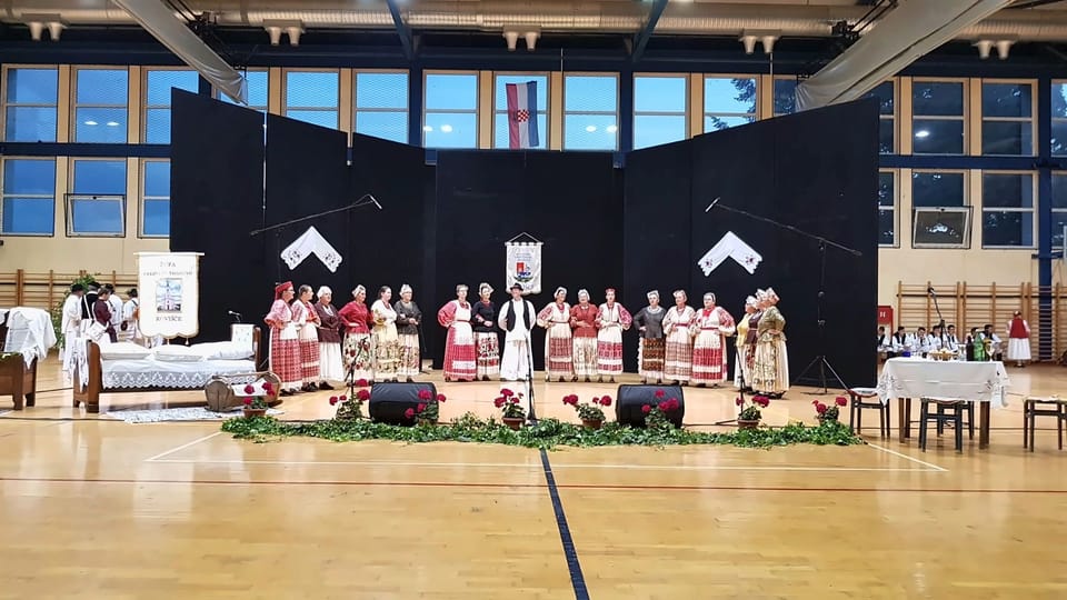 Pjevačka folklorna skupina u narodnim nošnjama na pozornici u sportskoj dvorani, ispred crnih scenskih zastora, s mikrofonima na stalcima i zvučnicima na podu.