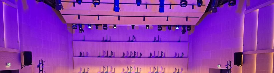 Koncertna dvorana u ljubičastom svjetlu: rasvjetne cijevi s reflektorima, stepenasti drveni zid s notama i zvučnici na bočnim zidovima.