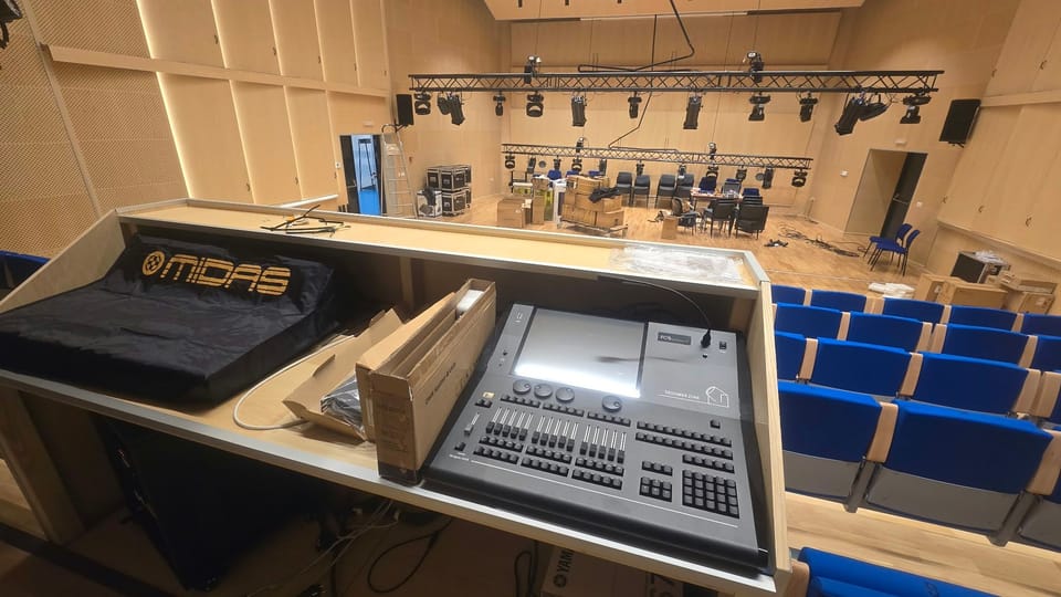 Pogled s kontrolnog pulta na stražnjem dijelu gledališta: rasvjetni pult FOS Designer 2048, mikseta MIDAS pod navlakom, plava sjedala i dvije truss linije s rasvjetnim tijelima iznad pozornice.