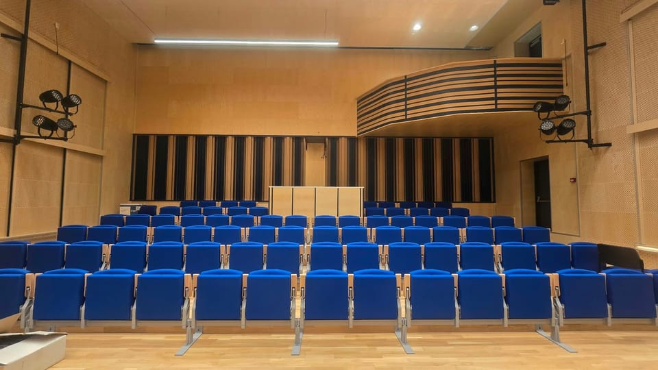 Pogled s pozornice prema gledalištu: redovi plavih sjedala, kontrolni pult u sredini zadnjeg reda, zakrivljena galerija s prugama i bočni stupovi s LED reflektorima.