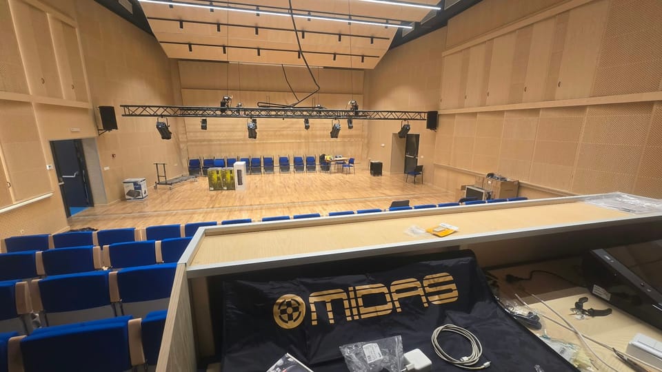 Pogled s kontrolnog pulta: mikseta MIDAS pod navlakom u prvom planu, truss s rasvjetnim tijelima, zvučnici na zidnim nosačima i drveni strop s tračnicama za rasvjetu.