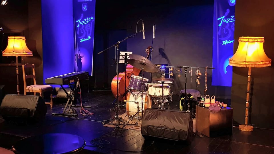 Pripremljena jazz pozornica s bubnjevima, kontrabasom, klavijaturom, mikrofonima i podnim monitorima pod plavim i toplim svjetlom.