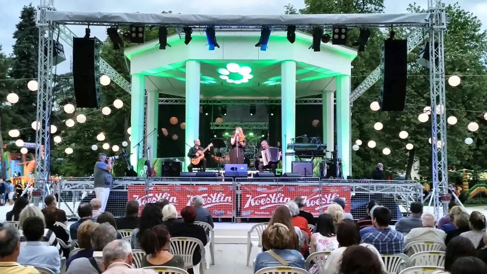 Koncert na otvorenom: paviljon osvijetljen zelenim svjetlom, truss tornjevi s ovješenim zvučnicima i rasvjetom, publika na stolicama.